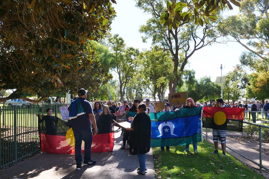 A small crowd, including people holding signs and Aboriginal and Torres Strait Islander flags, assemble in a park.