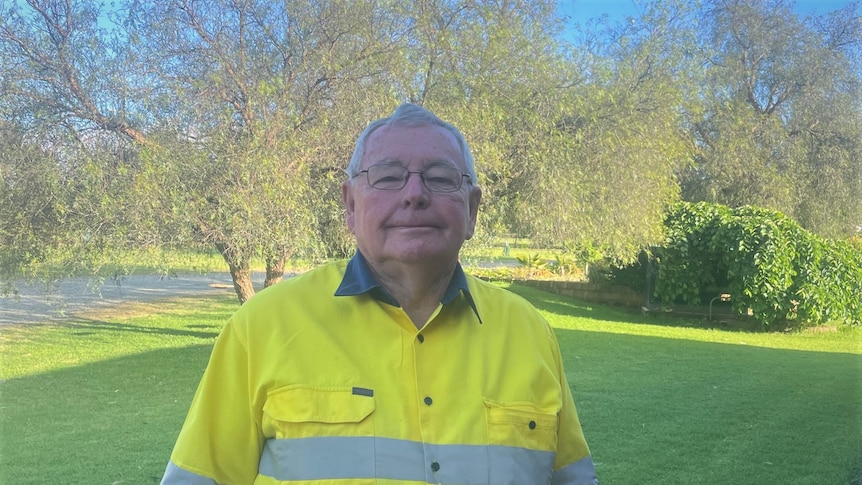 An smiling elderly man, grey hair, glasses, wears a high visibility top and stands in front of a green lawn.