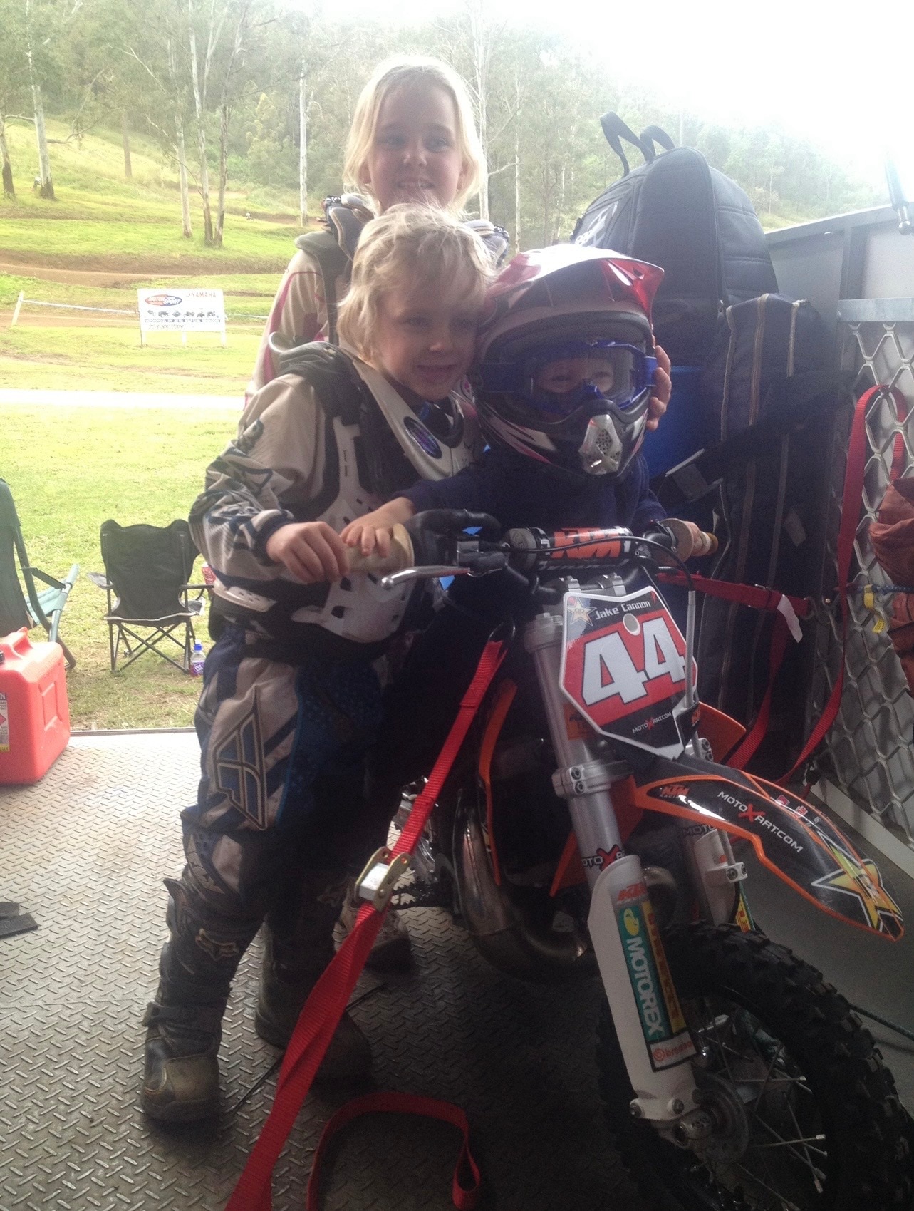 Três crianças vestidas com roupas de motocross cercam uma moto suja.