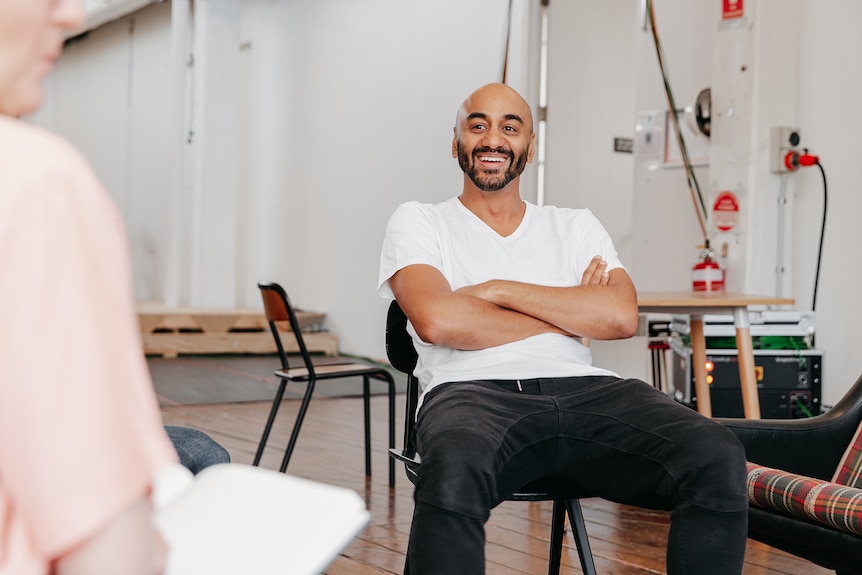 A Sri Lankan man in his early 40s, bald head, dark beard and white tshirt, sits on a chair with his arms crossed 