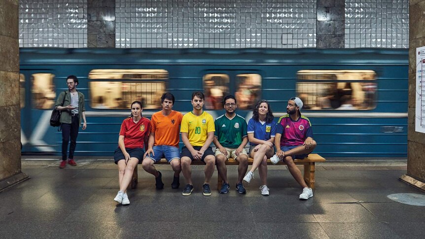 People wearing different coloured jerseys sit in a row in a subway station.