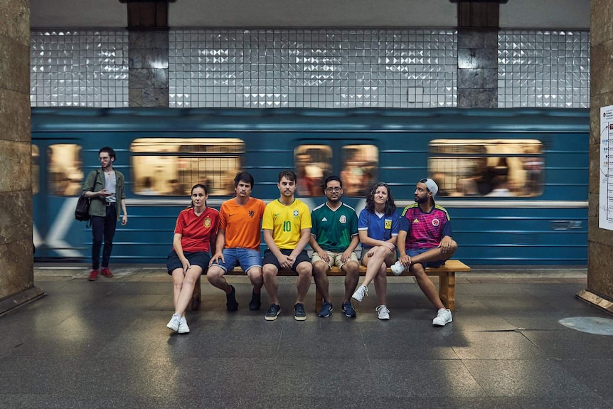 People wearing different coloured jerseys sit in a row in a subway station.