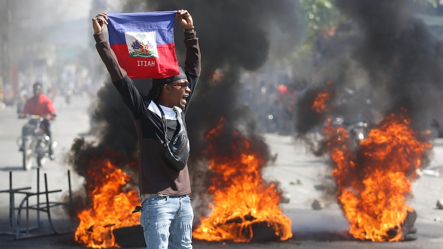 A demonstrator holds up an Haitian flag during protests. Three tyres are on fire behind him.