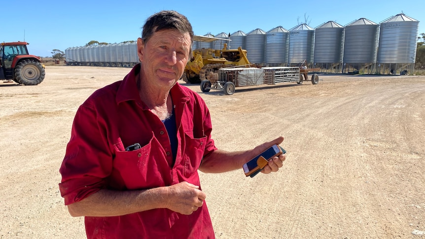 A man in a red shirt standing on a farm with a mobile phone in hand.