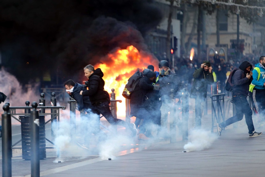 People run away from a burning car as tear canisters smoke in the foreground during clashes