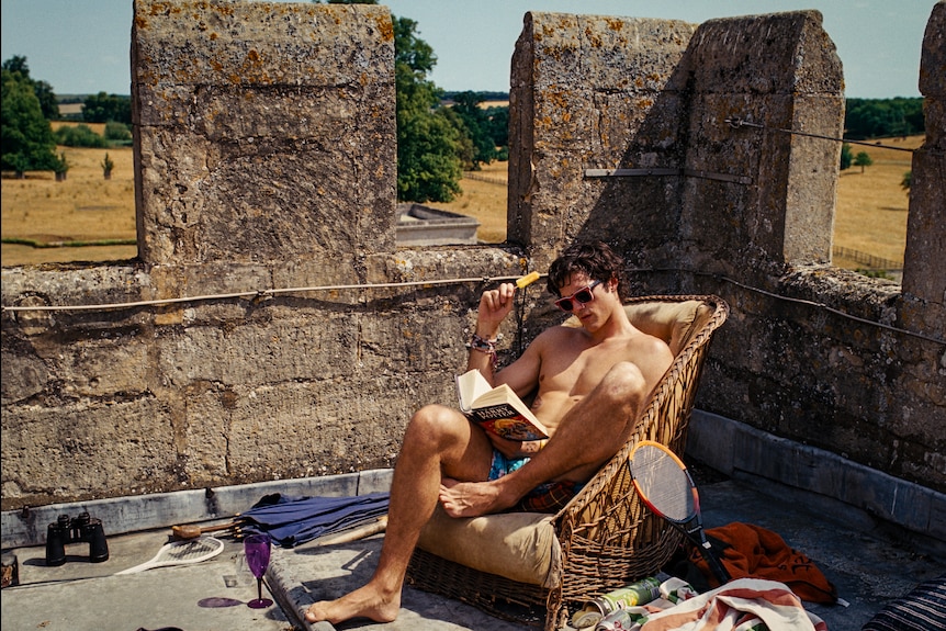 A man sitting outside reading a book and eating an icy pole.