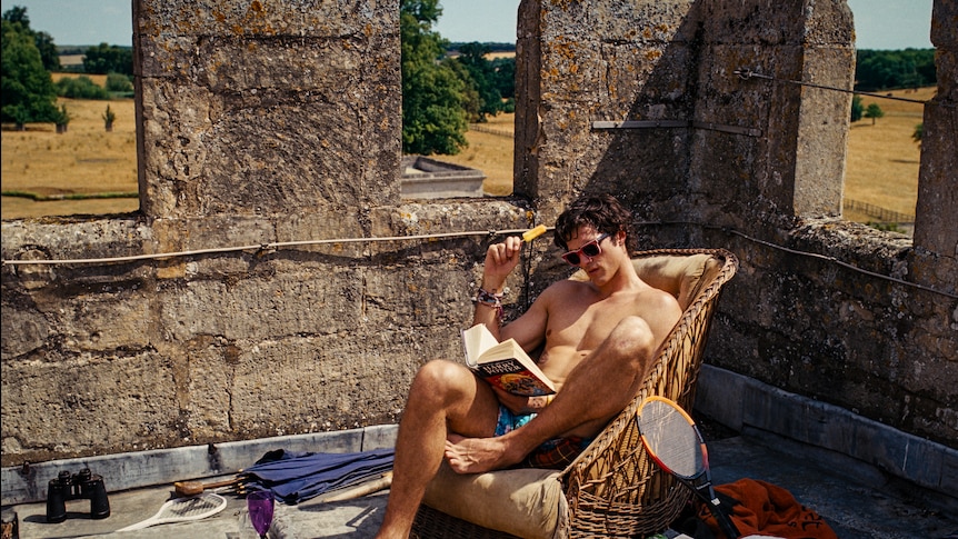 A shirtless young man reads a book on a rooftop with old stone barricades