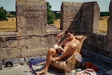 A shirtless young man reads a book on a rooftop with old stone barricades