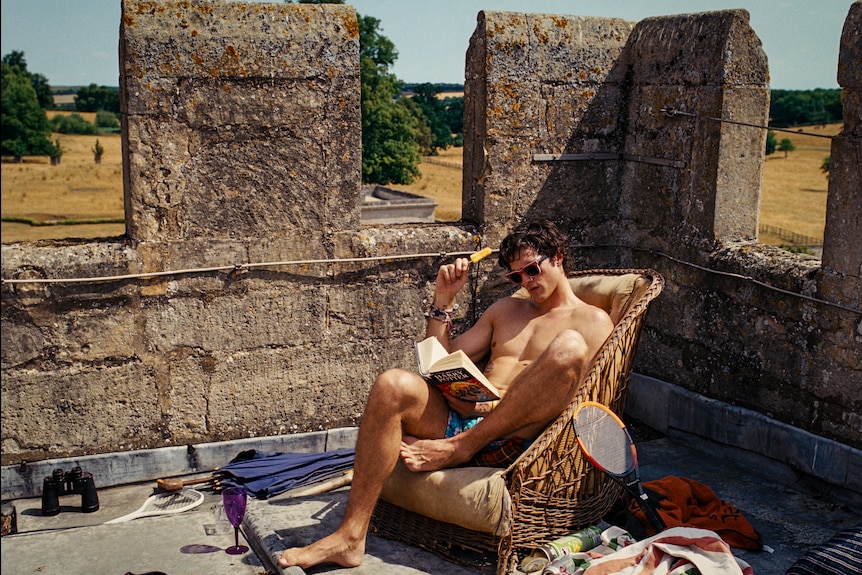 A shirtless young man reads a book on a rooftop with old stone barricades