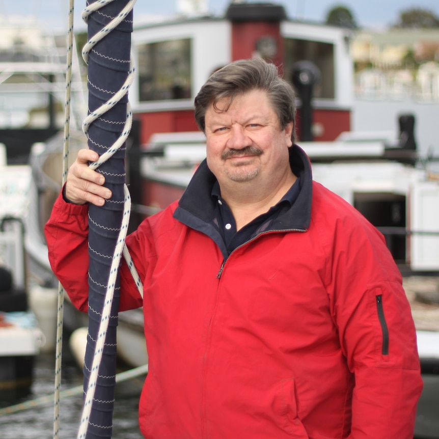 A man with brown hair, wearing a red outdoors jacket, standing next to a mast, with a harbour in the background.