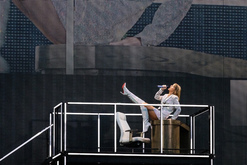 Taylor swift on a desk on stage with one leg in the air, wearing a sparkling silver jacket and singing into a microphone