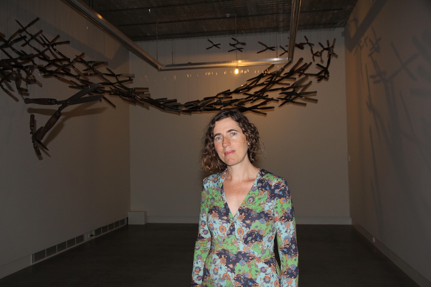 Tamara Dean stands inside a room wearing a floral dress with a wooden sculpture on the wall behind her.