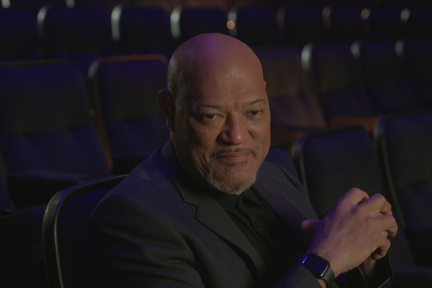Bald middle-aged black man with trim grey beard and moustache wears black shirt and suit and sits in seating bank of auditorium.