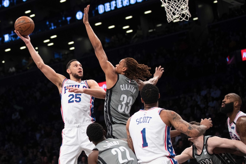 Ben Simmons holds a basketball in one hand and jumps in the air, looking at the basket