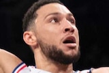 Ben Simmons holds a basketball in one hand and jumps in the air, looking at the basket