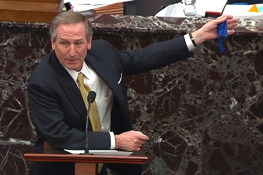 Michael van der Veen in a suit and tie holds out his left hand while standing at a lectern