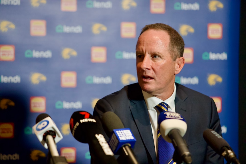 A man is speaking and looking to the side while sitting down with a row of microphones in front him at a press conference