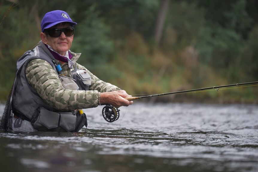 A woman fly fishing, wearing all the gear and standing in a river, looking to camera.