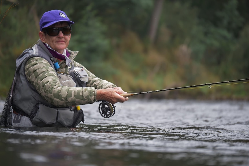 A woman fly fishing, wearing all the gear and standing in a river, looking to camera.