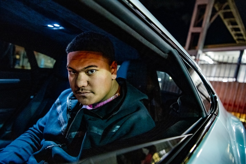 A young man sitting in the back seat of a car looks out the window at the camera at night.