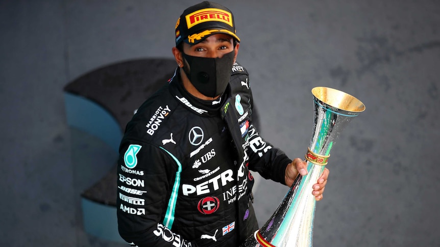 Lewis Hamilton looks up a camera, wearing a black mask, while holding a sliver trophy
