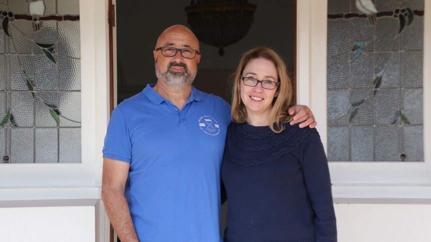 A man with his arm around a woman, both wearing glasses, outside the front door of their house.