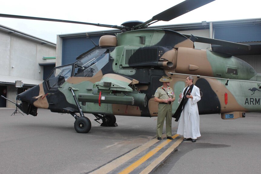 A military clergyman and a man in military dress stand in front of a helicopter.