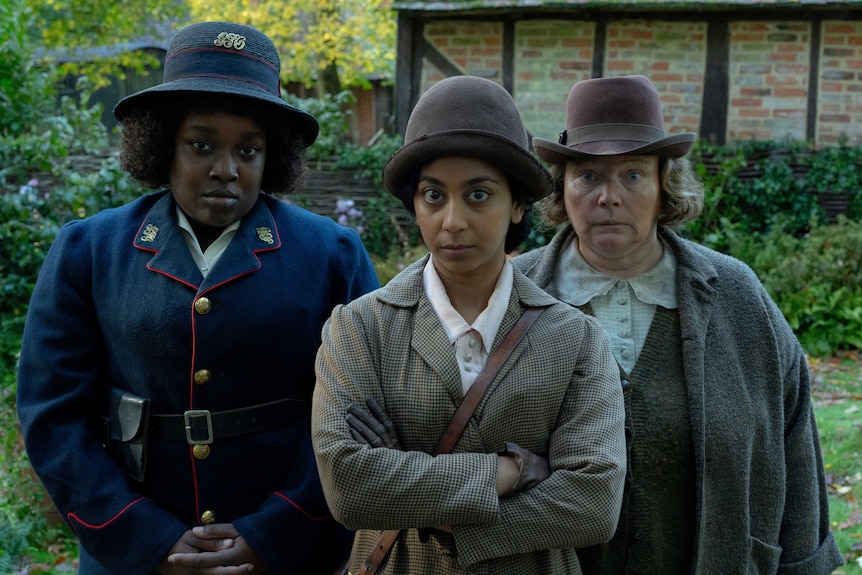 Three women, in 1920s uniforms, stand side by side at a doorstop in the British countryside.