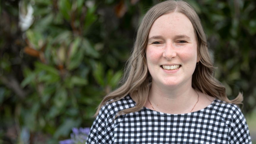 A young woman with light brown hair stands in a garden wearing a black and white checked top and smiles.