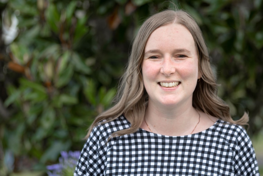 A young woman with light brown hair stands in a garden wearing a black and white checked top and smiles.