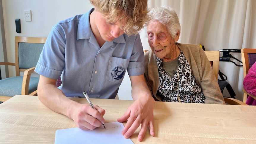 A teenage boy sitting with an elderly woman writes on a piece of paper as she looks on.