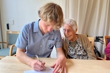 A teenage boy sitting with an elderly woman writes on a piece of paper as she looks on.