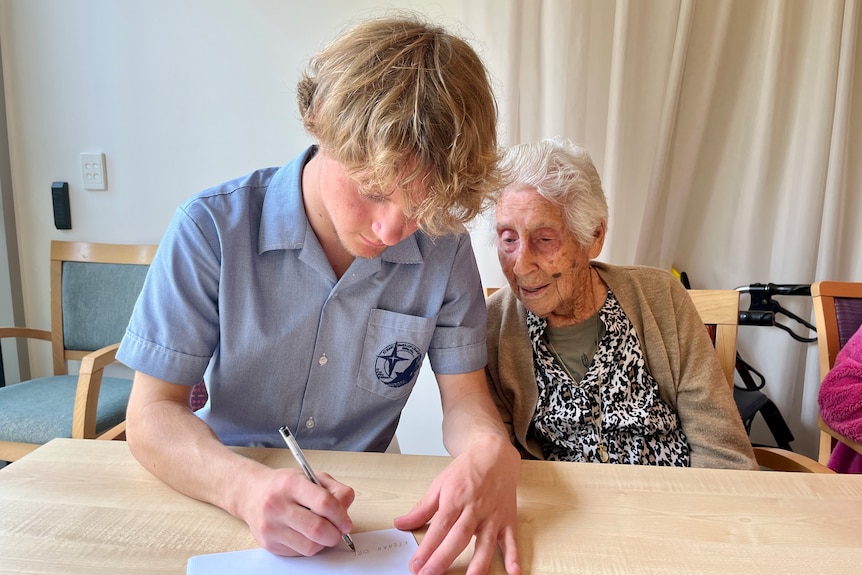 A teenage boy sitting with an elderly woman writes on a piece of paper as she looks on.