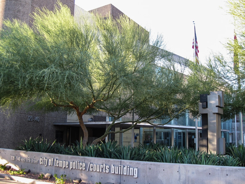 A police building exterior with a US flag flying above a concrete building with tress in the foreground.