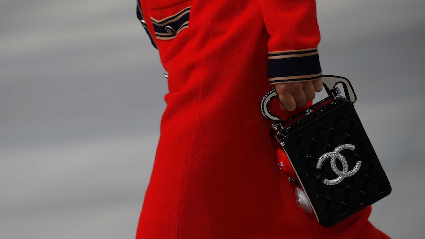 A model walks down the catwalk wearing a red suit and holding a black handbag