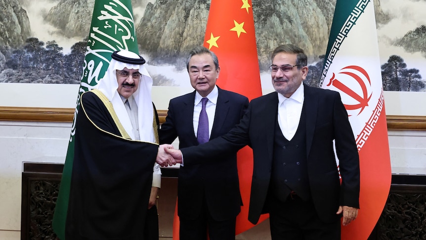 Three men stand together, with two either side shaking hands .