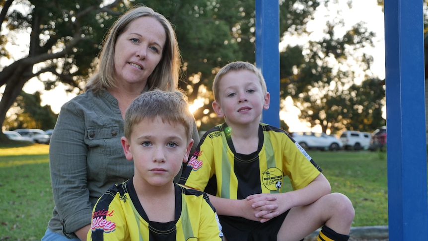 Sarah sits beside the soccer fields with her two sons Jasper and Charlie. The kids are wearing their footy uniforms. 