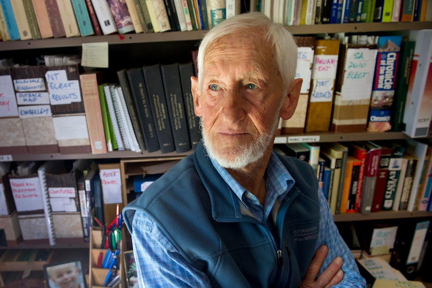 An elderly gentleman stands with his arms crossed in front of a bookshelf filled with books