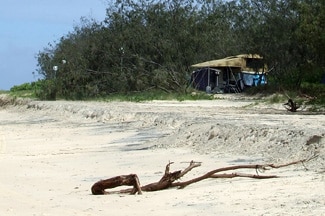 A log on a white sandy beach near trees and dunes and a campsite.