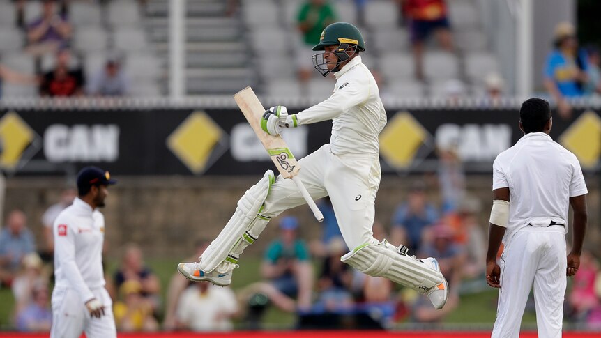Usman Khawaja jumps in the air with his legs apart, holding his bat in one hand