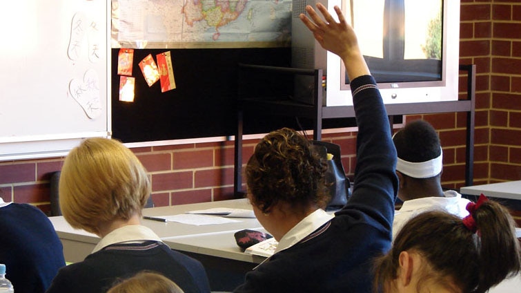 A student sits at a desk in a classroom with her hand in the air.