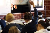 A shot of a secondary student sitting down and holding their hand up in the air in a classroom.