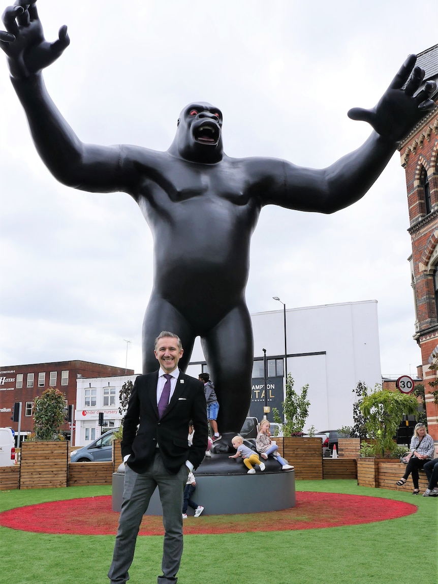 Un hombre vestido con jeans y un blazer negro se para frente a una estatua gigante de King Kong.