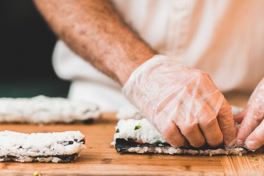 A man wearing gloves rolls rice with sheets of seaweed to make sushi.