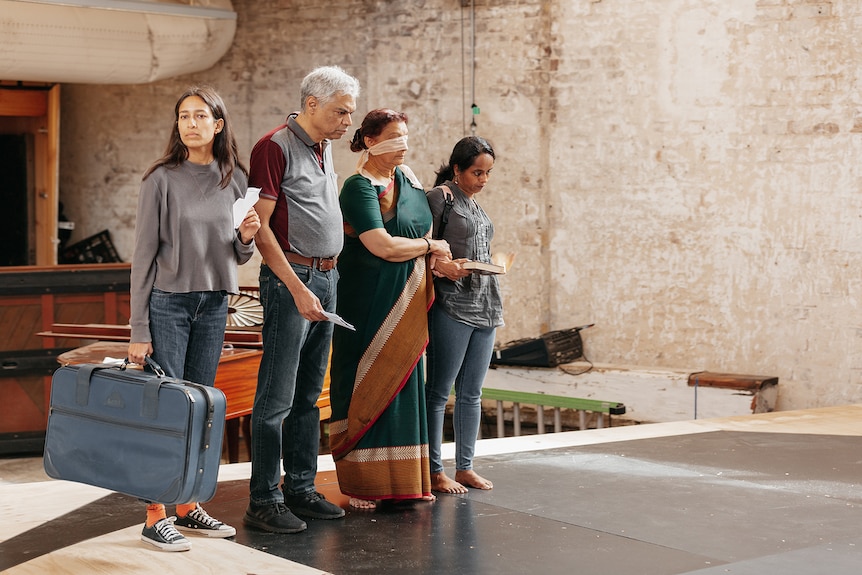 A Sri Lankan young woman, older man, older woman and a young woman stand in a row on a stage. The first woman holds a suitcase