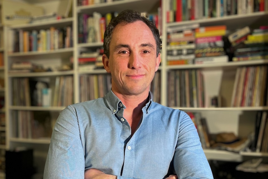 Simon McCrudden smiles for a portrait at home in front of a bookcase.