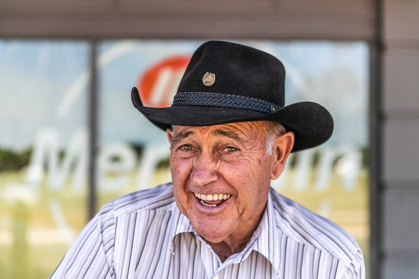 An old man with a cowboy hat on smiles at the camera.