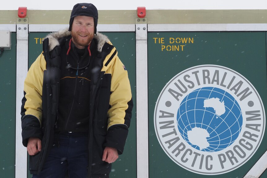A man in a beanie and a thick winter coat standing next to the logo of the Australian Antarctic Division