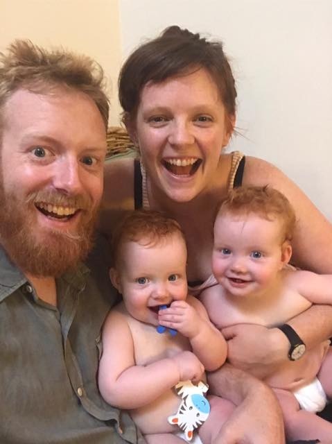 A smiling father and mother hold their twins, dressed in nappies.