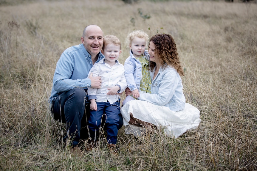 A lady and a man crouch down on grass while hugging two young children.